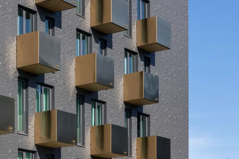 Cubos de balcón escalonados en tonos dorados sobre una estructura de fachada metálica mate de Sto.